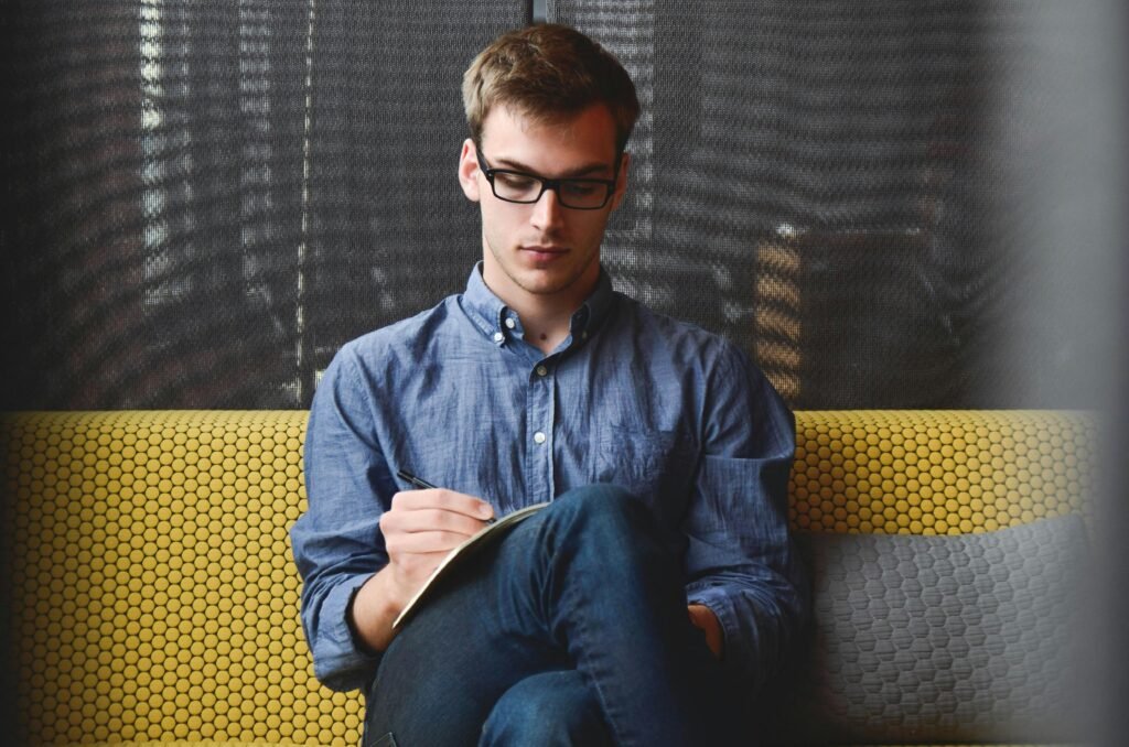 XL Digital : A young man in glasses writes in a notebook while sitting on a stylish couch indoors.