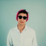 Fashionable young man wearing sunglasses and a pink beanie, set against a minimalist pastel background.